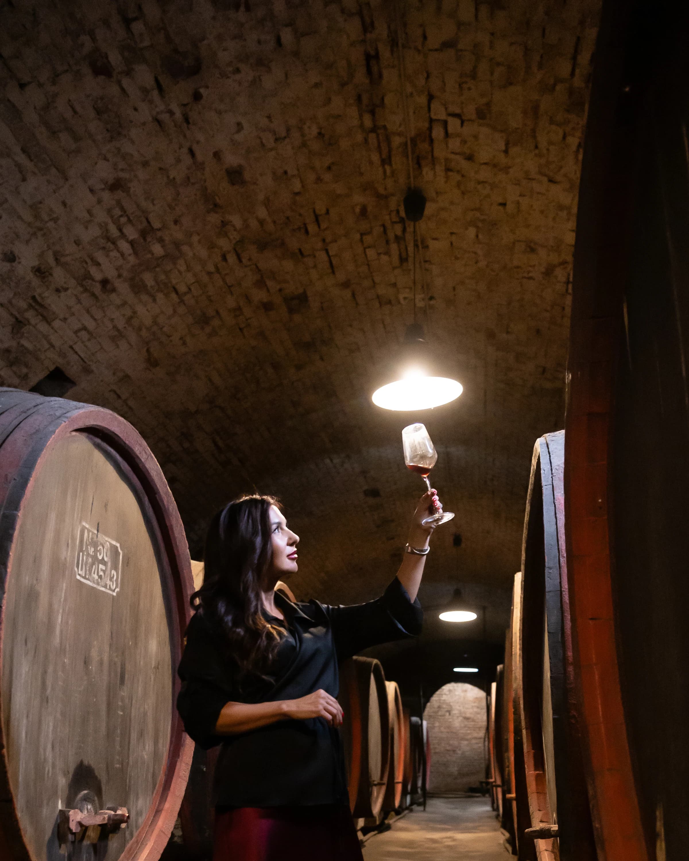 Roxana ridică un pahar de vin spre lumină, între butoaie mari de lemn
